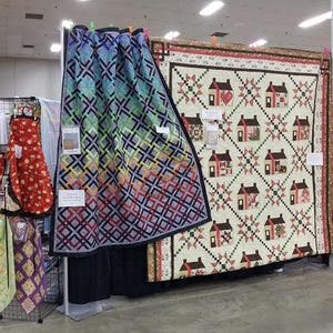 Peut inclure: Une exposition de courtepointes colorées avec des motifs géométriques et floraux. Les courtepointes sont accrochées à un mur dans une pièce avec un plafond blanc et des murs brun clair.