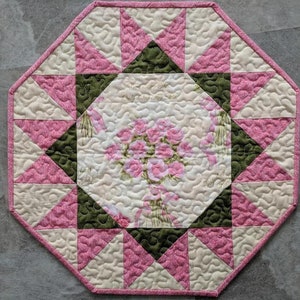 Op de afbeelding: Een achthoekige, gewatteerde placemat met een centraal bloemenboeketontwerp. De placemat heeft een roze, crème- en groen kleurenschema met een herhalend geometrisch patroon. De randen zijn afgewerkt met een roze bies.