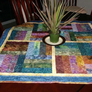 May include: A colorful patchwork quilt table runner with a blue border and a yellow inner border. A green plant in a brown pot sits on a white saucer in the center of the runner.