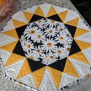 May include: A quilted, octagonal placemat with a daisy pattern in the center. The placemat features yellow, black, and white geometric shapes. The center has a black background with white daisies. The outer edge is white with a black and white border.