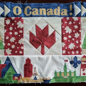 May include: A handmade quilt featuring a Canadian theme. The quilt is made of various fabrics and includes a red maple leaf, a green pine tree, and a red barn. The quilt also features the words "O Canada!" in gold lettering.