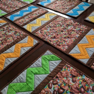 May include: Set of six colourful placemats with a chevron pattern. Each placemat features a brown border and a different coloured chevron pattern. The placemats are made of fabric and have a quilted texture.