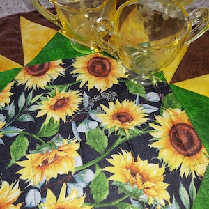 May include: A quilted table runner featuring a sunflower design on a black background. The runner is bordered by a patchwork design in brown, yellow and green. Two clear glass jugs are placed on top of the runner.