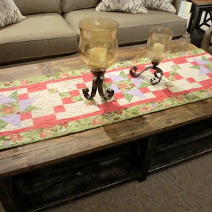 Może przedstawiać: Tkany bieżnik stołowy z kwiatowym wzorem w kolorze różowym, zielonym i białym. Bieżnik leży na drewnianym stoliku kawowym z dwoma świecznikami.