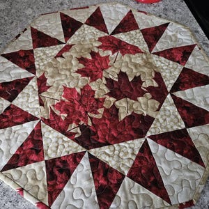 May include: Octagonal table runner with a quilted design featuring a central motif of red maple leaves on a cream background. The runner is surrounded by a repeating pattern of red and cream triangles.
