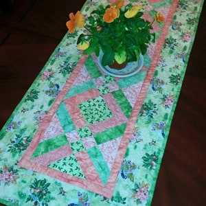 May include: A green and pink quilted table runner with a floral pattern and a green border. The runner is decorated with a small potted plant with orange flowers.