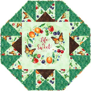 Könnte beinhalten: Eine grüne und braune Patchwork-Decke mit einem zentralen Quadrat, das einen Kranz aus Früchten, Blumen und Schmetterlingen zeigt. Der Kranz trägt den Text "Life is Sweet" in der Mitte.