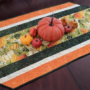 Pode incluir: Um corredor de mesa acolchoado com um design listrado verde, branco e laranja. O corredor apresenta uma estampa de abóbora e abóbora e é decorado com pequenas abóboras decorativas.