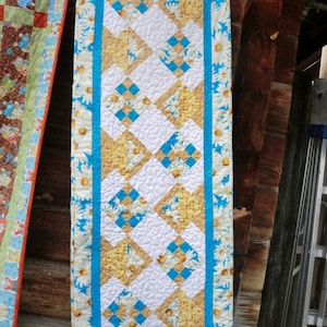 Könnte beinhalten: Ein blauer und weißer Tischläufer mit einem Gänseblümchen-Druckrand und einem Patchwork-Design in der Mitte. Das Patchwork-Design zeigt gelbe, weiße und blaue Quadrate.