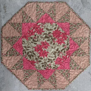 May include: Octagonal table runner with a starburst design in shades of pink and green. The centre features a floral pattern with poinsettia flowers and holly leaves.
