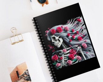 May include: A spiral-bound notebook with a black cover featuring a skeleton with flowing gray hair and a crown of red roses. The skeleton's rib cage is adorned with more roses. The notebook is open on a white surface.