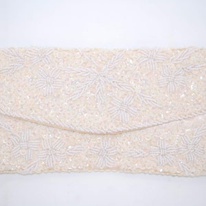Op de afbeelding: Ivoor kleurige, met kralen bezette clutch met een overslag. De clutch is bedekt met kleine, iriserende pailletten en heeft een bloemmotief gemaakt met witte kralen. Het ontwerp is rechthoekig.