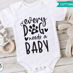 Op de afbeelding: Witte baby romper met zwarte tekst die "every dog needs a baby" luidt. De tekst bevat een pootprint en een hart. Een knuffeldier is op de achtergrond.