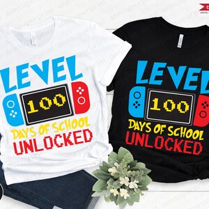 Op de afbeelding: Twee T-shirts met een videogameontwerp. Het witte T-shirt heeft blauwe en rode accenten, terwijl het zwarte T-shirt rode en blauwe accenten heeft. Beide T-shirts tonen de tekst "Level 100 Dagen School Ontgrendeld".