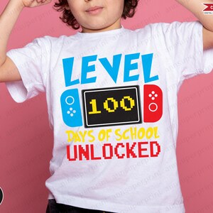 Op de afbeelding: Een wit T-shirt met een blauw, geel en rood grafisch ontwerp. De afbeelding toont een videogamecontroller met de tekst "LEVEL 100 DAYS OF SCHOOL UNLOCKED".