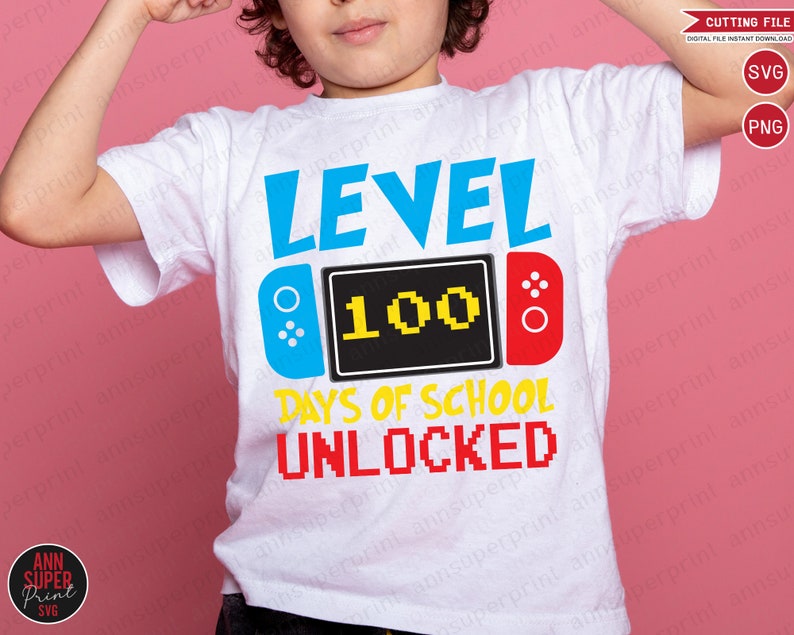 Op de afbeelding: Een wit T-shirt met een blauw, geel en rood grafisch ontwerp. De afbeelding toont een videogamecontroller met de tekst "LEVEL 100 DAYS OF SCHOOL UNLOCKED".