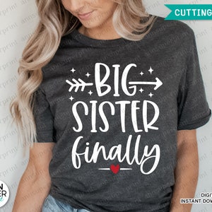 May include: A dark gray t-shirt with white text that reads "BIG SISTER finally" with a small red heart. The text is in a handwritten font and includes a small arrow pointing to the right.