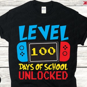 Op de afbeelding: Zwart T-shirt met een blauwe en rode videogamecontroller-afbeelding. De afbeelding zegt "Level 100 dagen school ontgrendeld".