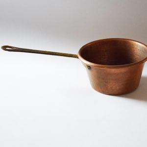 Könnte beinhalten: Ein Kupfer-Kochtopf mit einem langen, dünnen Griff. Der Topf hat einen abgerundeten Körper und einen leicht ausgestellten Rand. Der Griff ist mit Nieten befestigt. Das Kupfer hat eine warme, rötlich-braune Farbe.