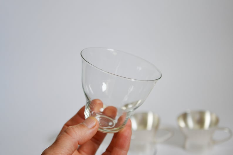 Peut inclure: Un verre en verre transparent avec un bord large et &eacute;vas&eacute; et une base &eacute;troite. Le verre est tenu dans une main, et deux autres verres sont flous en arri&egrave;re-plan.