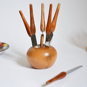 Może przedstawiać: Drewniany stojak na noże w kształcie jabłka z pięcioma nożami z drewnianymi rękojeściami. Noże są ułożone w kształcie gwiazdy, a rękojeści skierowane są do góry. Stojak wykonany jest z jasnobrązowego drewna i ma gładkie wykończenie.