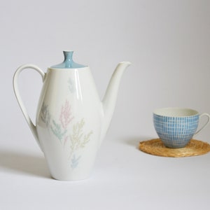 Op de afbeelding: Een witte theepot van keramiek met een blauw deksel en een bloemenmotief in roze, groene en blauwe tinten. De theepot heeft een tuit en een handvat.