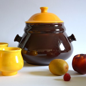 May include: A brown ceramic pot with a yellow lid and four yellow ceramic cups. The pot has two handles on the sides. There is a kiwi fruit, a peach, and a raspberry on the table.