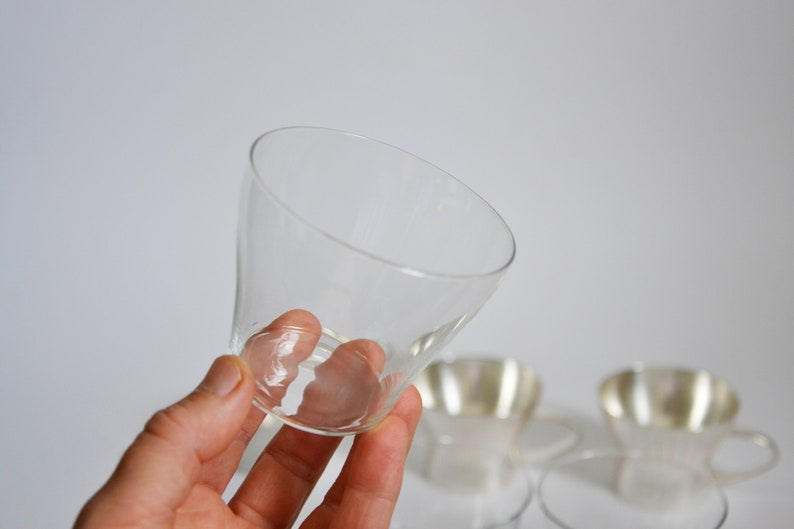 Peut inclure: Une tasse en verre transparent avec un bord large et &eacute;vas&eacute; et une base &eacute;troite. La tasse est tenue dans une main, et il y a deux autres tasses en arri&egrave;re-plan.
