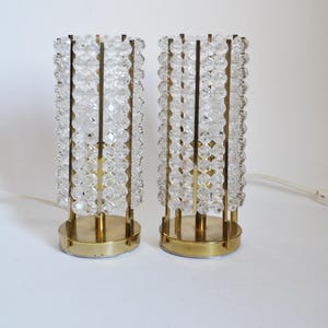 Op de afbeelding: Twee cilindrische tafellampen met een goudkleurige voet en frame. De lampen zijn versierd met heldere, facetgeslepen kristallen kralen, wat een sprankelend effect creëert. Elke lamp heeft een wit elektrisch snoer en een schakelaar.