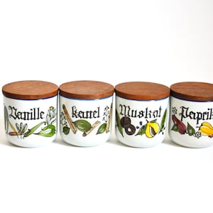 Könnte beinhalten: Vier weiße Keramik-Gewürzgläser mit Holzdeckeln. Jedes Glas zeigt ein anderes Gewürz: Vanille, Zimt, Muskatnuss und Paprika. Die Gläser sind in einer Reihe von links nach rechts angeordnet.