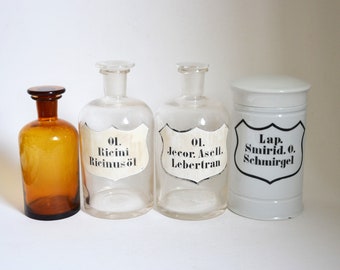 May include: Three vintage glass bottles with labels and a white ceramic jar with a lid. The bottles are clear glass and the jar is white with black lettering. The labels on the bottles read "01. Ricini Ricinus öl", "01. Jecor: Asell. Lebertran", and "Lap. Smirid. 0. Schmirgel".