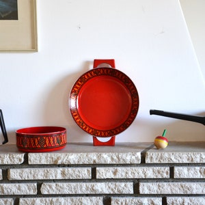 Könnte beinhalten: Ein Set aus rotem Emaille-Kochgeschirr mit schwarzen Griffen und einem geometrischen Muster. Das Set enthält einen Topf, einen Topf und eine Bratpfanne. Das Kochgeschirr steht auf einer Backsteinmauer.