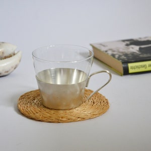 Peut inclure: Une tasse en verre avec une bande argent&eacute;e autour du milieu, pos&eacute;e sur un dessous de tasse en osier. La tasse a une anse et est vide.