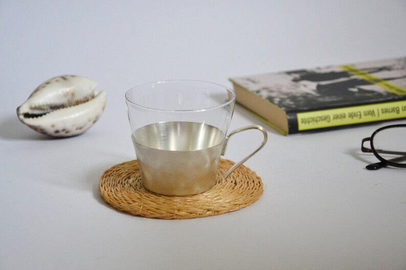 Peut inclure: Une tasse en verre avec une bande argent&eacute;e autour du milieu, pos&eacute;e sur un dessous de tasse en osier. La tasse a une anse et est vide.