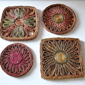 Könnte beinhalten: Vier gewebte Untersetzer in verschiedenen Formen: zwei quadratische und zwei runde. Die Untersetzer weisen florale Designs mit Farben wie Braun, Grün, Rot und Gelb auf. Sie bestehen aus einem natürlichen Fasermaterial.