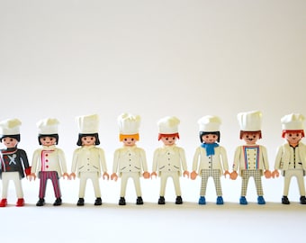 May include: A row of eight white plastic figurines dressed as chefs, each wearing a white chef's hat and a different colored outfit. The chefs are standing in a line, facing forward.