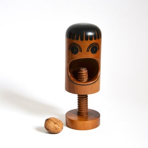 Puede incluir: Un cascanueces de madera con forma de cara con una sonrisa y ojos pintados de negro. El cascanueces tiene una base de madera marrón y un tornillo de metal.