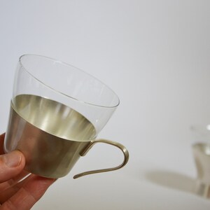 Peut inclure: Une tasse en verre transparent avec une bande en m&eacute;tal argent&eacute; autour du haut et une poign&eacute;e en m&eacute;tal argent&eacute;. La tasse est tenue dans une main.