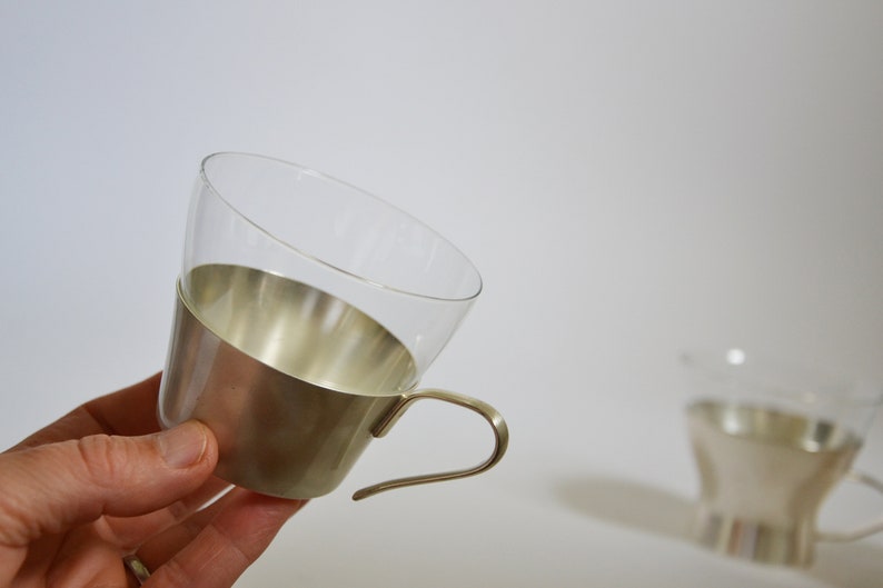 Peut inclure: Une tasse en verre transparent avec une bande en m&eacute;tal argent&eacute; autour du haut et une poign&eacute;e en m&eacute;tal argent&eacute;. La tasse est tenue dans une main.