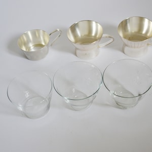 Peut inclure: Six tasses en argent et en verre, trois de chaque, dispos&eacute;es en rang&eacute;e sur une surface blanche. Les tasses en argent ont des anses et sont l&eacute;g&egrave;rement &eacute;vas&eacute;es en haut. Les tasses en verre sont transparentes et ont une base l&eacute;g&egrave;rement plus large.
