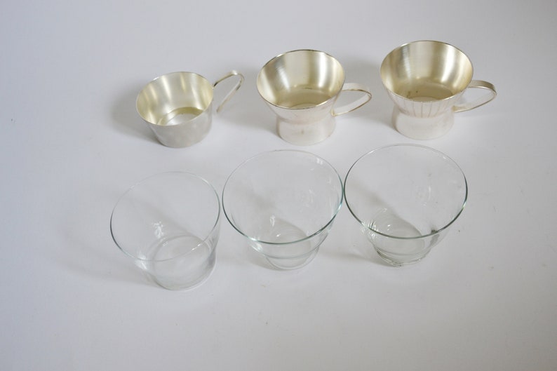Peut inclure: Six tasses en argent et en verre, trois de chaque, dispos&eacute;es en rang&eacute;e sur une surface blanche. Les tasses en argent ont des anses et sont l&eacute;g&egrave;rement &eacute;vas&eacute;es en haut. Les tasses en verre sont transparentes et ont une base l&eacute;g&egrave;rement plus large.