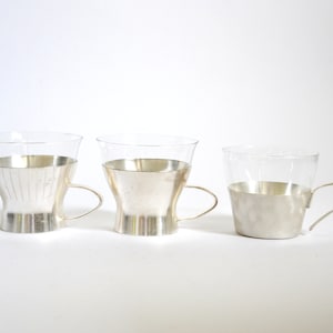 Peut inclure: Trois tasses en verre avec des bords argent&eacute;s et des poign&eacute;es. Les tasses sont dispos&eacute;es en rang&eacute;e de gauche &agrave; droite, la plus grande tasse &agrave; gauche et la plus petite tasse &agrave; droite.