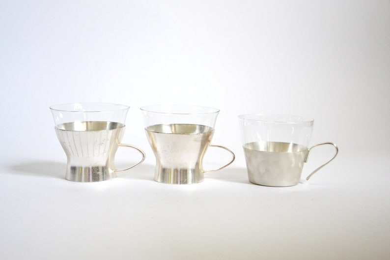 Peut inclure: Trois tasses en verre avec des bords argent&eacute;s et des poign&eacute;es. Les tasses sont dispos&eacute;es en rang&eacute;e de gauche &agrave; droite, la plus grande tasse &agrave; gauche et la plus petite tasse &agrave; droite.