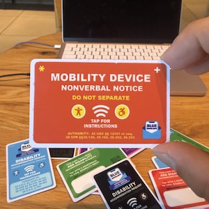 May include: A rectangular orange card with white text reading MOBILITY DEVICE NONVERBAL NOTICE DO NOT SEPARATE. It features yellow icons of a person and a wheelchair, a Wi-Fi symbol, and the text TAP FOR INSTRUCTIONS. Includes legal text and a Blue Envelope logo.