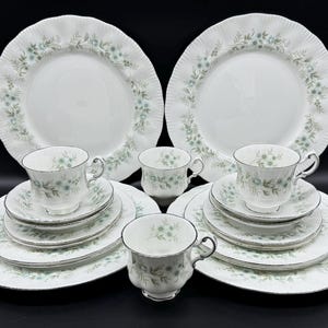 May include: A set of white china dinnerware with a delicate floral pattern in shades of green and blue. The set includes a large dinner plate, a side plate, a teacup, and a saucer.