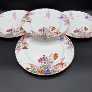 May include: Four white ceramic plates with scalloped edges. Each plate features a floral design with pink, blue, orange, and red flowers, accented with gold trim. The plates are arranged on a dark surface.