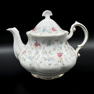 Peut inclure: Une théière en porcelaine blanche avec un motif floral. La théière est ornée d'une bordure dorée autour du couvercle et de la base. Le motif floral présente des fleurs roses, bleues et jaunes.