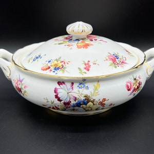 Puede incluir: Un plato de servir de porcelana blanca con tapa, con un patrón floral en tonos de rosa, azul, amarillo y verde. El plato tiene un borde dorado y un pomo dorado en la tapa.