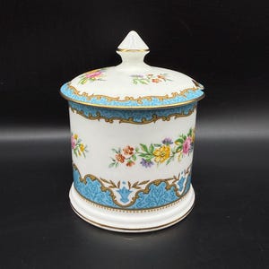 Puede incluir: Un tarro de cerámica blanca con tapa, decorado con un patrón floral azul y dorado. El tarro tiene un diseño floral con flores rosas, amarillas y azules.