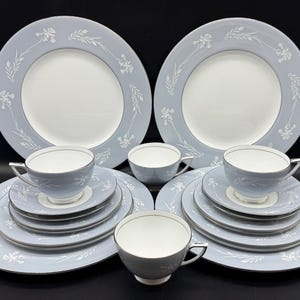 May include: A full dinner service with light blue plates, saucers, and cups, decorated with a white floral pattern and silver edging. The set includes large dinner plates, smaller plates, and teacups, all arranged on a black surface.